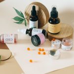 Frascos de óleo, cápsulas e outros produtos de canabidiol dispostos sobre fundo neutro, acompanhados de folhas de cannabis.