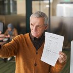 Eleitor idoso segura uma cédula e usa o smartphone dentro de uma cabine de votação.